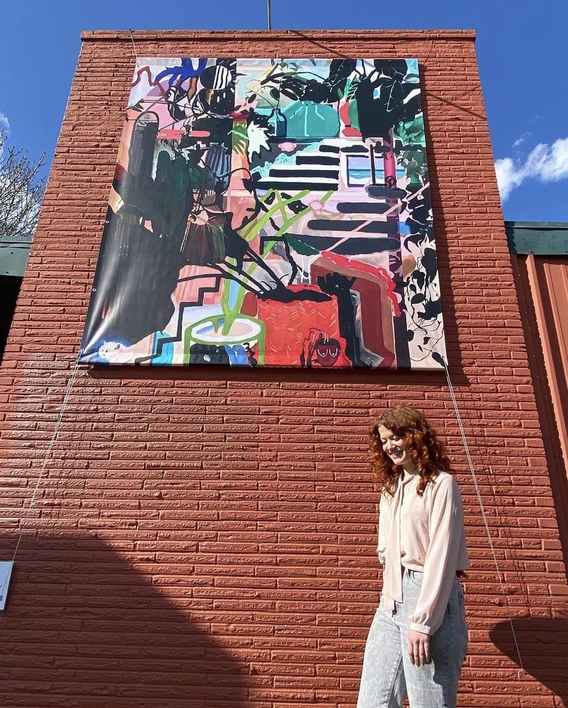 Emily Wise posant devant l'une de ses œuvres exposée en grand sur un mur de briques rouge